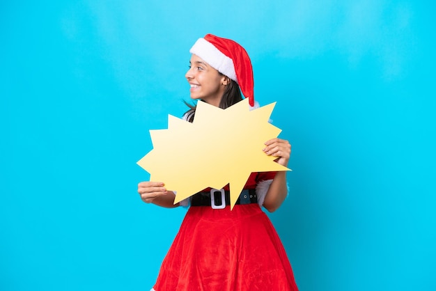 Photo young hispanic woman dressed as mama noel isolated on blue background holding an empty speech bubble