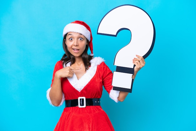 Young hispanic woman dressed as mama noel isolated on blue background holding a question mark icon with surprised expression