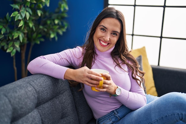 Young hispanic woman drinking coffee sitting on sofa at home