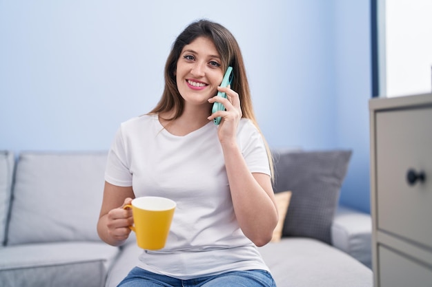 Молодая латиноамериканка пьет кофе и разговаривает по смартфону дома