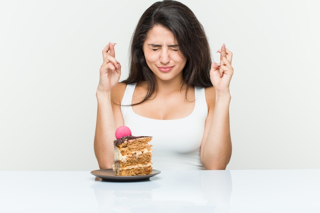 Young hispanic woman eating a cake crossing fingers for having luck