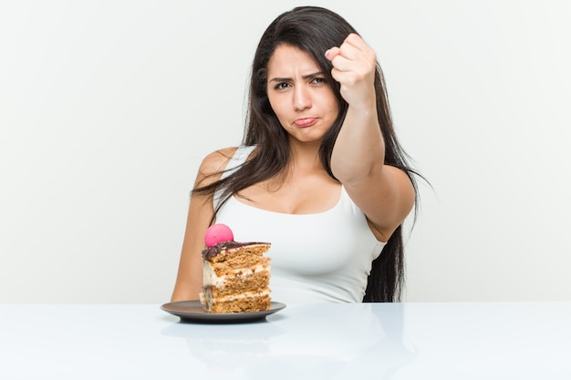 Young hispanic woman eating a cake showing fist to camera, aggressive facial expression.