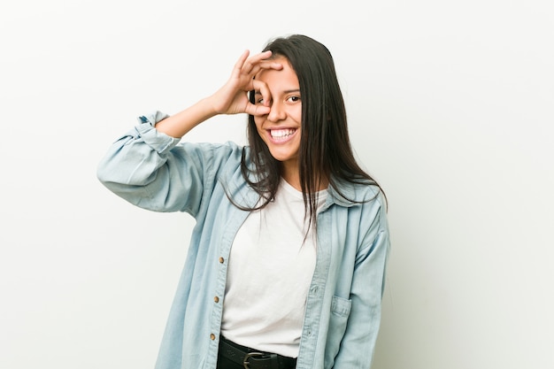 Young hispanic woman excited keeping ok gesture on eye.