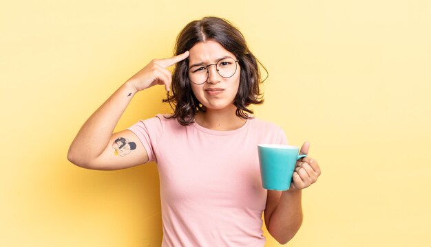 Young hispanic woman feeling confused and puzzled, showing you are insane. coffee mug concept