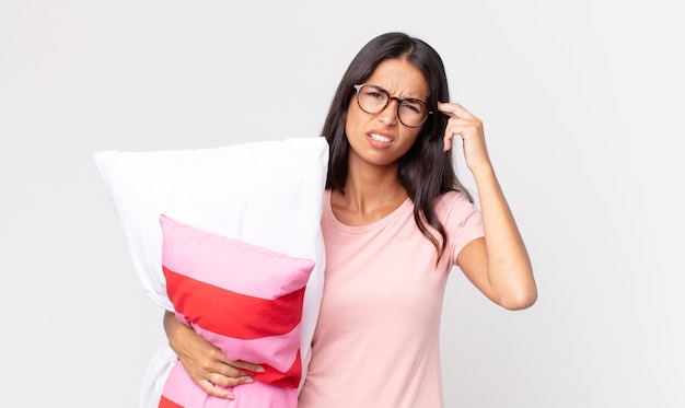 Young hispanic woman feeling confused and puzzled, showing you are insane wearing pajamas and holding a pillow