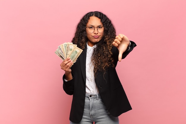 Young hispanic woman feeling cross, angry, annoyed, disappointed or displeased, showing thumbs down with a serious look. dollar banknotes concept