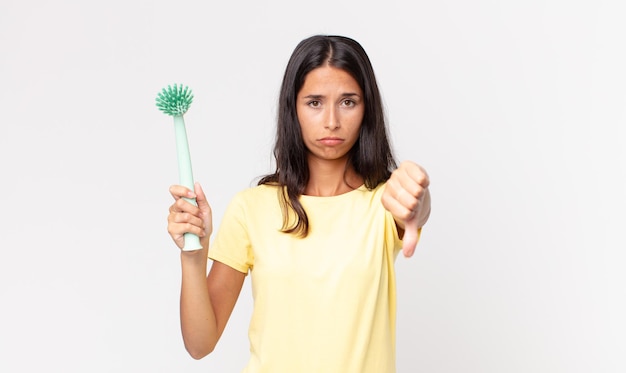 Young hispanic woman feeling cross,showing thumbs down and holding a dish cleaning brush