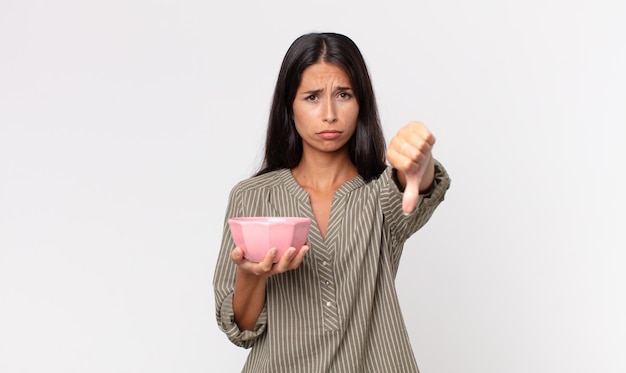 Young hispanic woman feeling cross,showing thumbs down and holding an empty bowl or pot