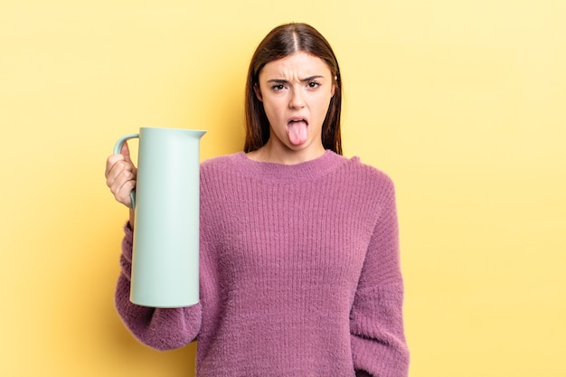 Young hispanic woman feeling disgusted and irritated and tongue out. coffee thermos concept