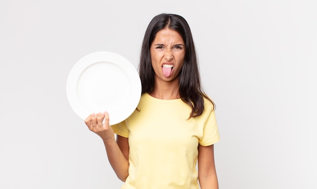 Young hispanic woman feeling disgusted and irritated and tongue out and holding an empty plate