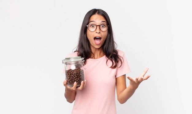 young hispanic woman feeling extremely shocked and surprised and holding a coffee beans bottle