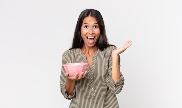 Young hispanic woman feeling happy and astonished at something unbelievable and holding an empty bowl or pot