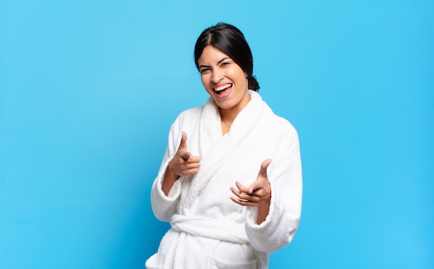 Young hispanic woman feeling happy, cool, satisfied, relaxed and successful, pointing at camera, choosing you. bathrobe