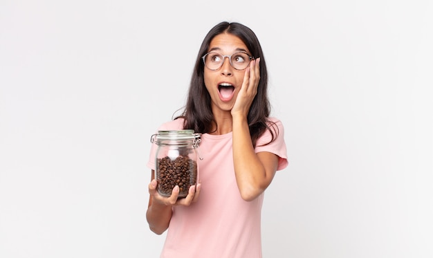 Young hispanic woman feeling happy, excited and surprised and holding a coffee beans bottle