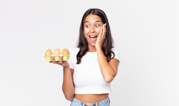 Young hispanic woman feeling happy, excited and surprised and holding an eggs box