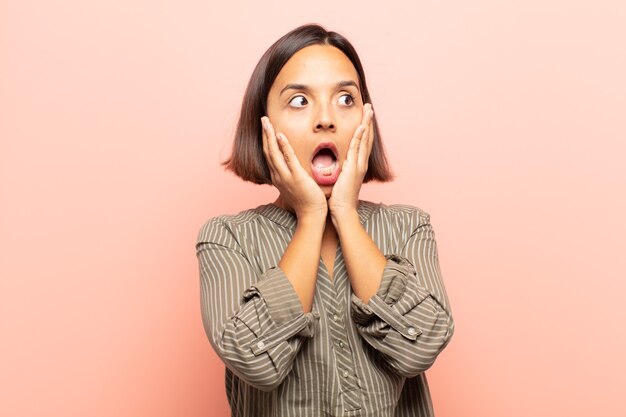 Young hispanic woman feeling happy, excited and surprised, looking to the side with both hands on face