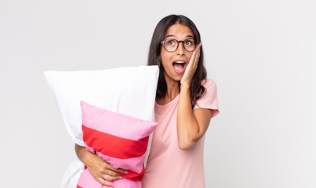 Young hispanic woman feeling happy, excited and surprised wearing pajamas and holding a pillow