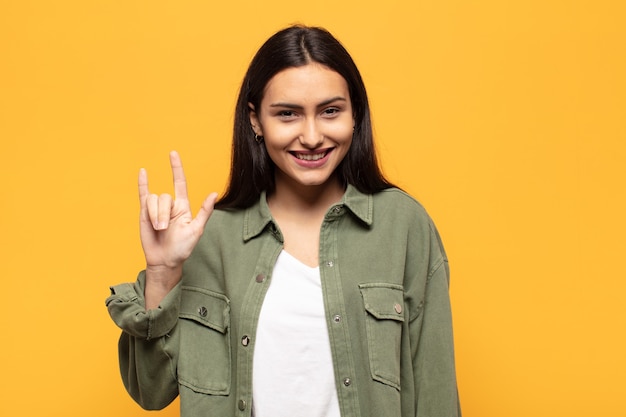 Young hispanic woman feeling happy, fun, confident, positive and rebellious, making rock or heavy metal sign with hand