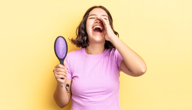 Young hispanic woman feeling happy,giving a big shout out with hands next to mouth. hairbrush concept