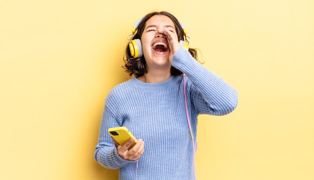 Young hispanic woman feeling happy,giving a big shout out with hands next to mouth. headphones and smartphone concept