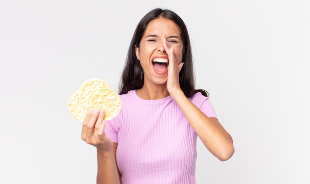 young hispanic woman feeling happy,giving a big shout out with hands next to mouth and holding a rice cookie. diet concept