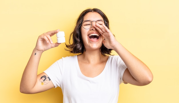 Young hispanic woman feeling happy,giving a big shout out with\
hands next to mouth. illness pills concept