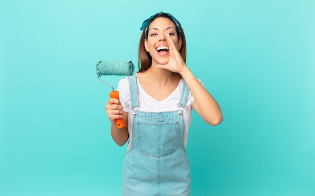 Young hispanic woman feeling happy,giving a big shout out with hands next to mouth and painting a wall