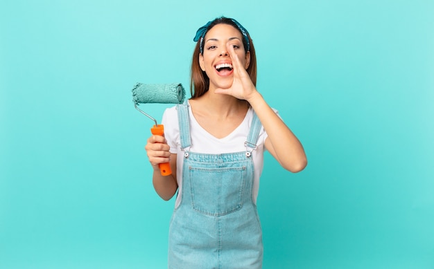 Young hispanic woman feeling happy,giving a big shout out with\
hands next to mouth and painting a wall