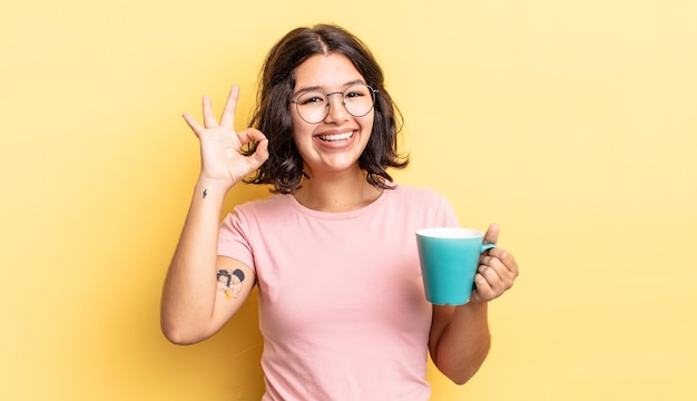 Young hispanic woman feeling happy, showing approval with okay gesture. coffee mug concept