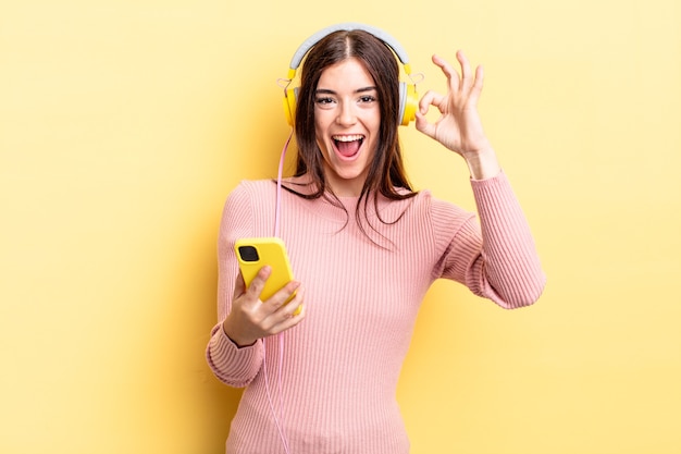 young hispanic woman feeling happy, showing approval with okay gesture. headphones and telephone concept