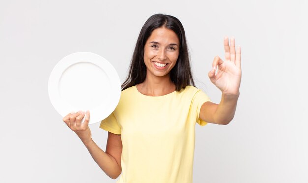 Young hispanic woman feeling happy, showing approval with okay gesture and holding an empty plate