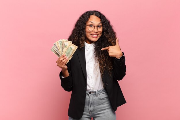 Young hispanic woman feeling happy, surprised and proud, pointing to self with an excited, amazed look. dollar banknotes concept