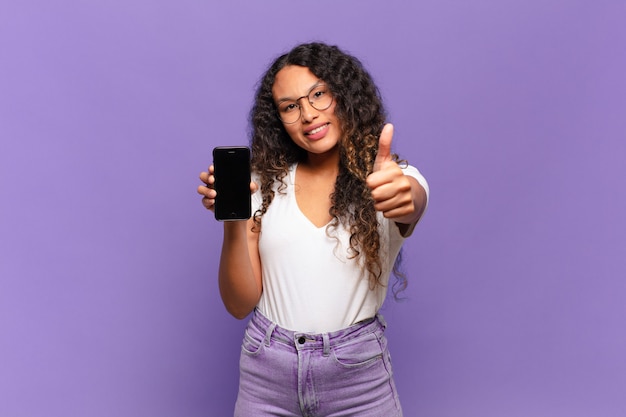 Young hispanic woman feeling proud, carefree, confident and happy, smiling positively with thumbs up. smart phone concept