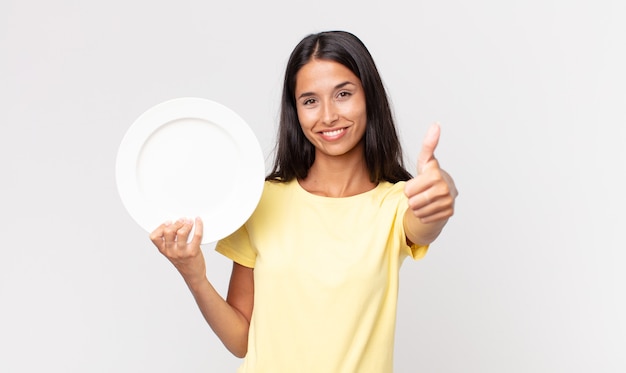 Young hispanic woman feeling proud,smiling positively with thumbs up and holding an empty plate
