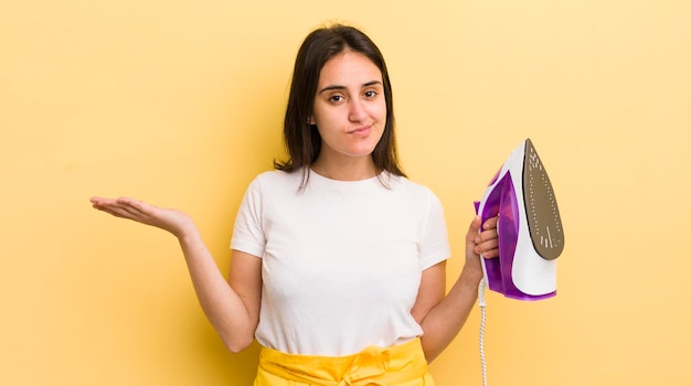 Young hispanic woman feeling puzzled and confused and doubting clothes iron concept