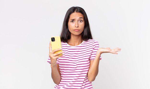 Young hispanic woman feeling puzzled and confused and doubting and holding a smartphone