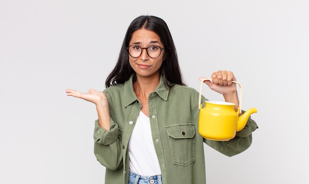 Young hispanic woman feeling puzzled and confused and doubting and holding a tea pot