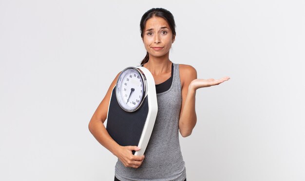 困惑し、混乱し、体重計を疑って保持していると感じている若いヒスパニック系女性。ダイエットコンセプト