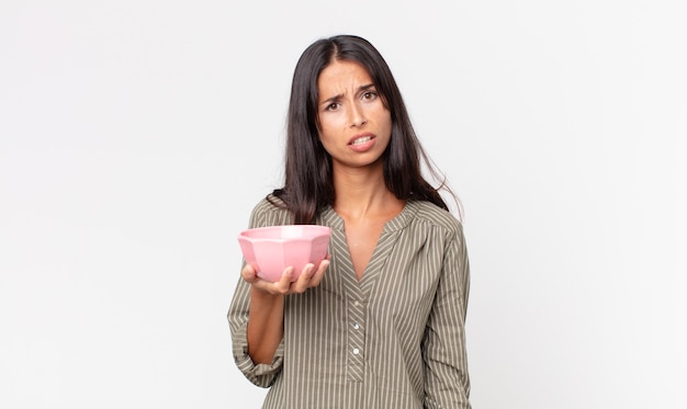 Young hispanic woman feeling puzzled and confused and holding an empty bowl or pot