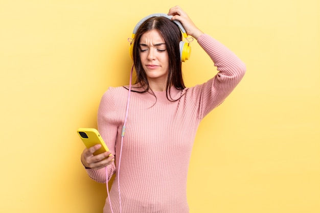 Young hispanic woman feeling puzzled and confused, scratching head. headphones and telephone concept