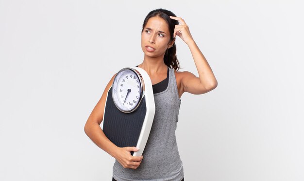 Young hispanic woman feeling puzzled and confused, scratching head and holding a weight scale. diet concept