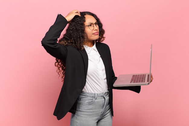 Young hispanic woman feeling puzzled and confused, scratching head and looking to the side. laptop concept