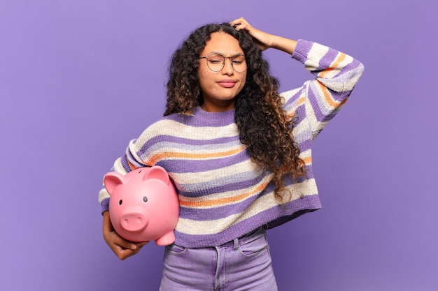 Young hispanic woman feeling puzzled and confused, scratching head and looking to the side. piggy bank concept