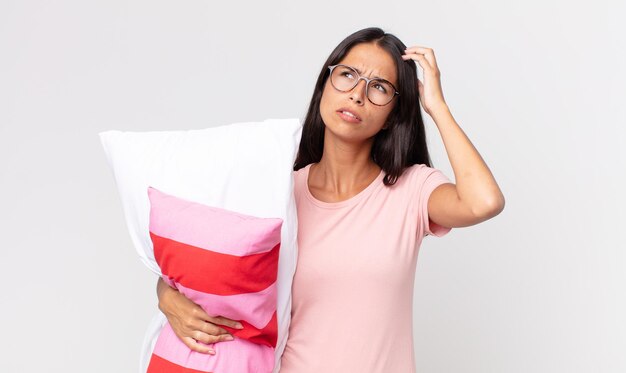 Young hispanic woman feeling puzzled and confused, scratching head wearing pajamas and holding a pillow