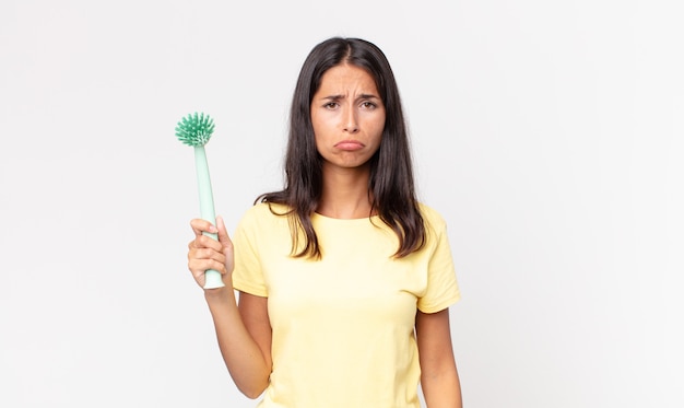 Giovane donna ispanica che si sente triste e piagnucolona con uno sguardo infelice e piange e tiene in mano una spazzola per la pulizia dei piatti
