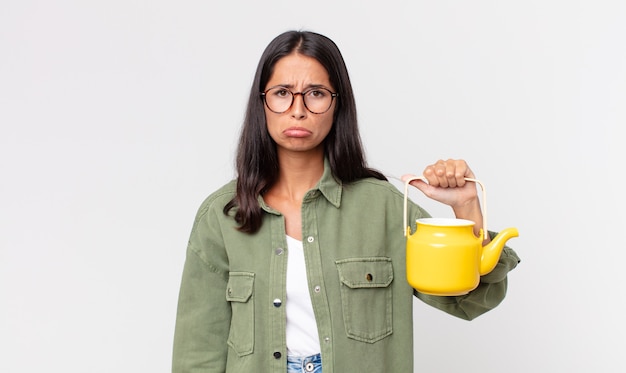 Young hispanic woman feeling sad and whiney with an unhappy look and crying and holding a tea pot