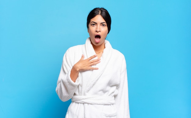 Young hispanic woman feeling shocked, astonished and surprised, with hand on chest and open mouth, saying who, me?. bathrobe concept