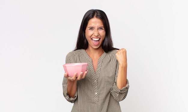 Young hispanic woman feeling shocked,laughing and celebrating success and holding an empty bowl or pot