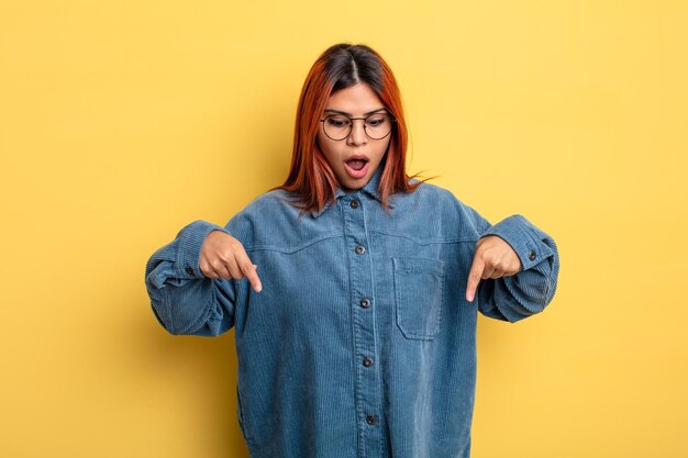 Young hispanic woman feeling shocked, open-mouthed and amazed, looking and pointing downwards in disbelief and surprise