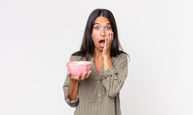 Young hispanic woman feeling shocked and scared and holding an empty bowl or pot